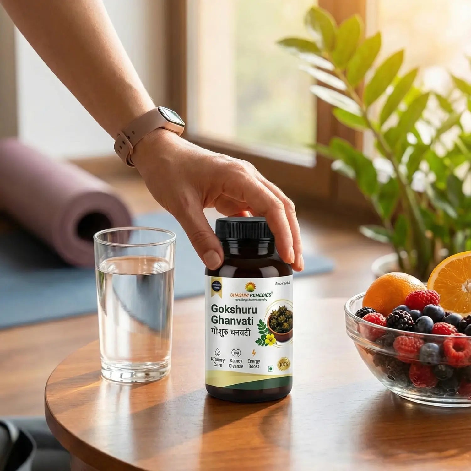 Hand holding a bottle of 'Gokshuru Ghanvati' with a glass of water and fruit bowl on a wooden table.